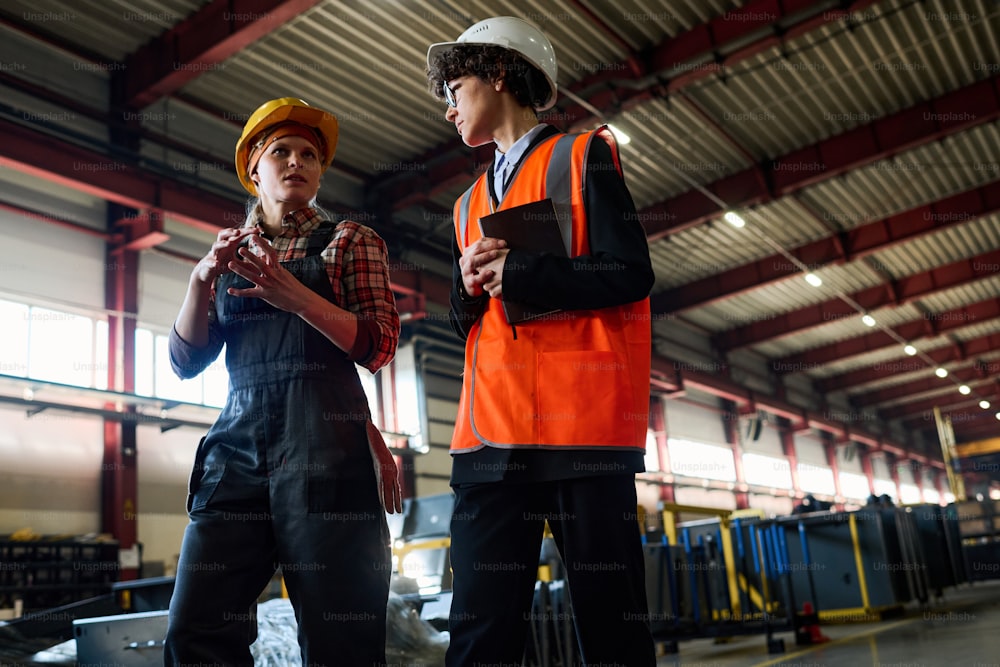 Young female engineer in hardhat and coveralls explaining her opinion to colleague during discussion of working moments on the move
