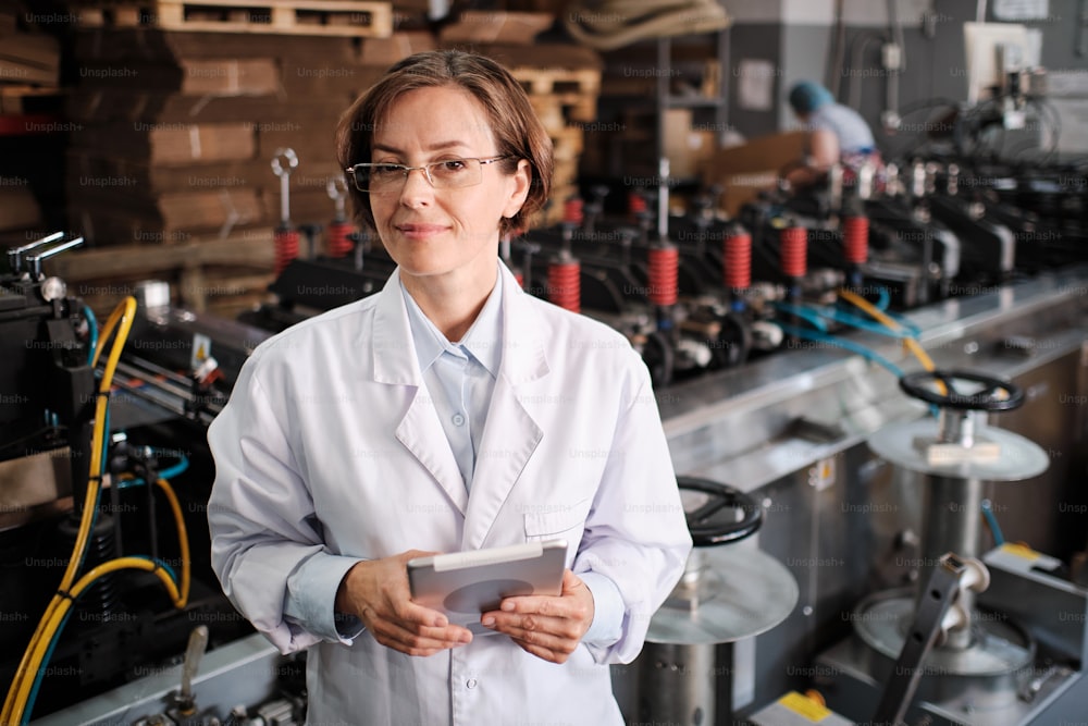 Portrait of female factory worker wearing glasses and white robe at polymer plastic manufacturing, using tablet for quality control and logistic purposes, checking machines and mechanisms