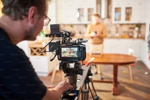 Videógrafo do sexo masculino jovem de pé ao lado da câmera de vídeo enquanto filma anúncio de culinária ou masterclass na cozinha