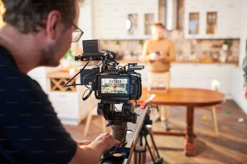 Young male videographer standing by video camera while shooting advertisement of cooking or masterclass in the kitchen
