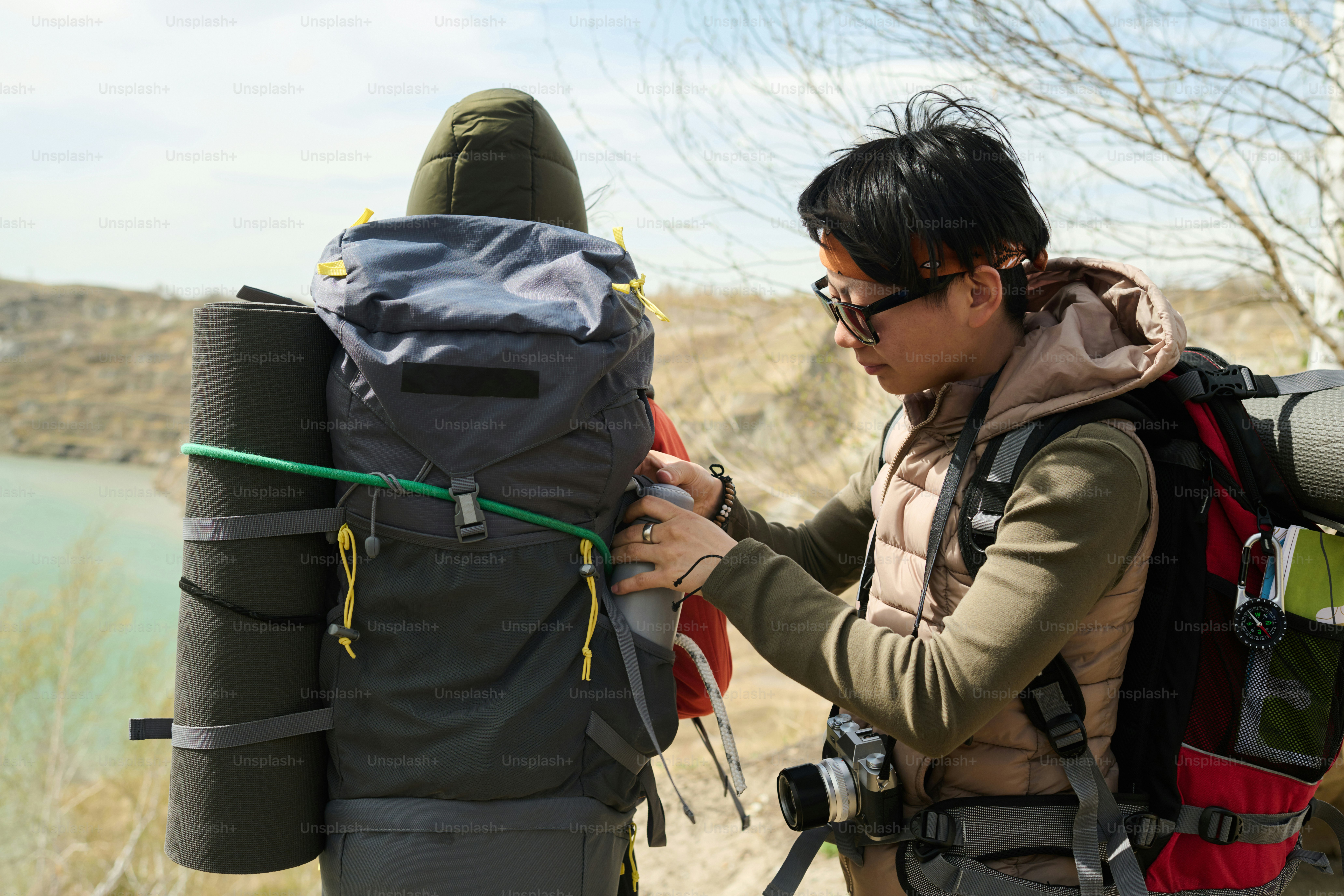 Young Asian female tourist taking out vacuum flask with hot tea from her friends backpack while hiking somewhere around quarry lake