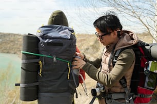 Jeune touriste asiatique sortant une bouteille isotherme avec du thé chaud du sac à dos de ses amis lors d’une randonnée quelque part autour du lac de la carrière