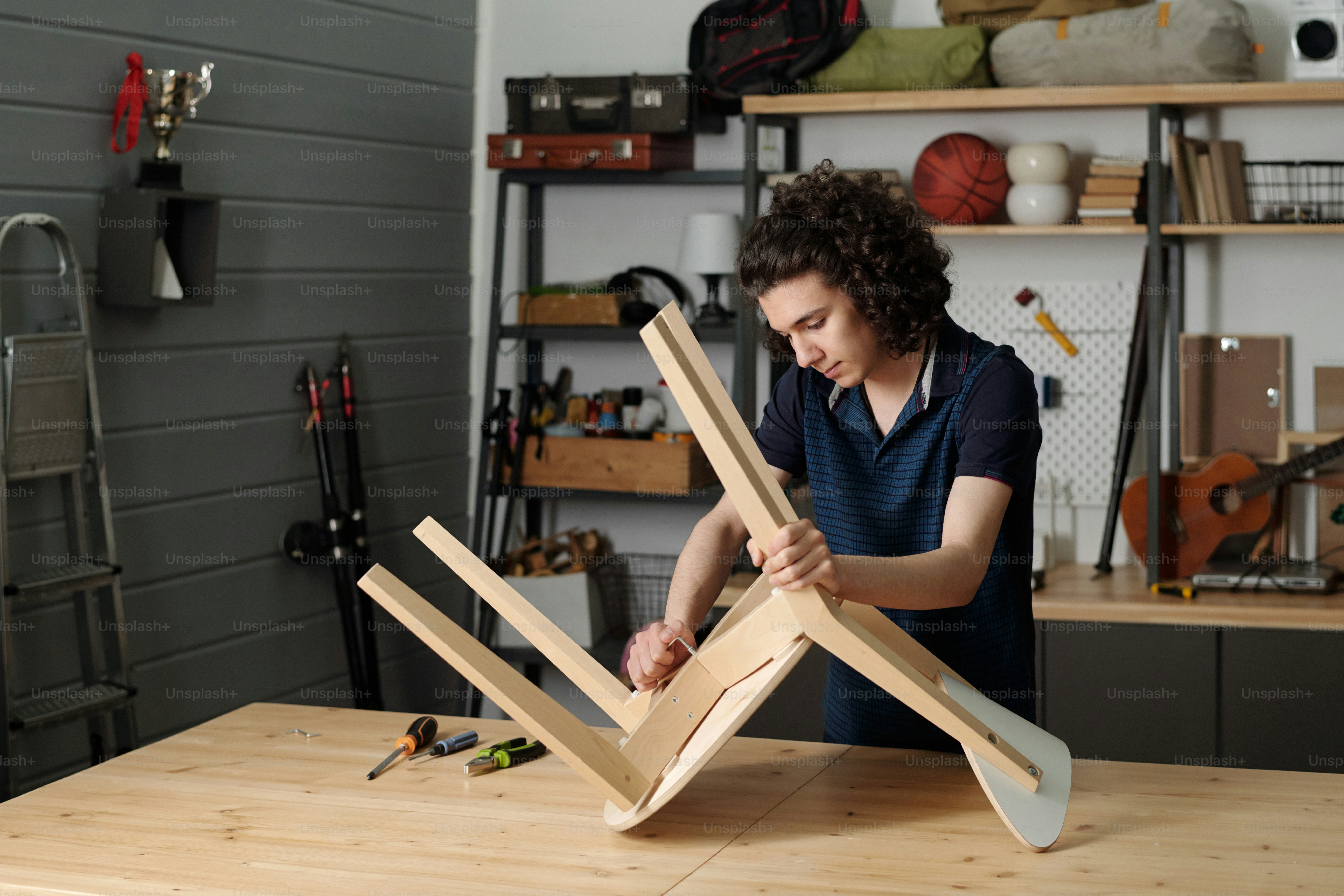 Youthful serious guy with handtool fixing part of wooden chair while standing by by table against shelves with domestic stuff in garage