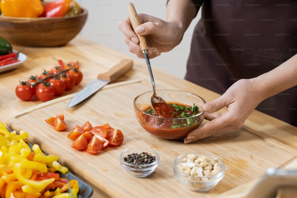 Hände der Frau, die hausgemachten Ketchup mit Petersilienblättern in Glasschüssel mischen, während vegetarisches Essen in der Küche gekocht wird