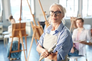 Mature confident teacher with paintbrushes looking at camera during lesson of painting in school of arts