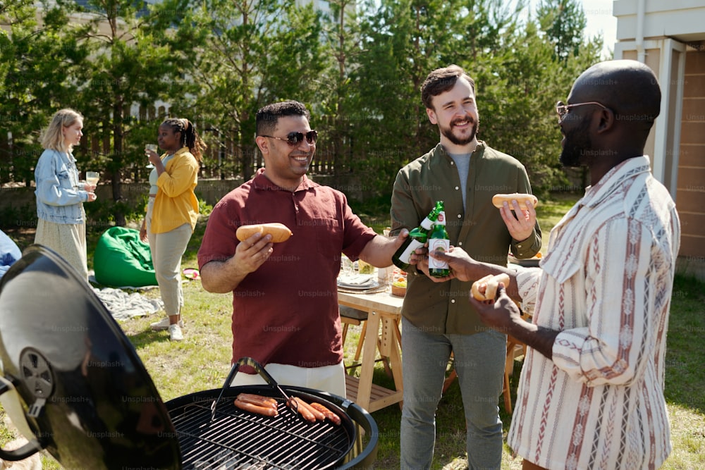 Gruppe junger fröhlicher interkultureller Männer, die während einer Party im Freien mit Bierflaschen gegen den servierten Tisch klirren und zwei Mädchen