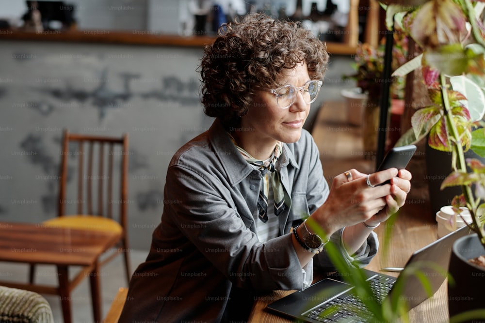 Junge Geschäftsfrau oder Freiberuflerin in Freizeitkleidung und Brille sitzt vor dem Laptop im Café und scrollt auf dem Handy oder SMS