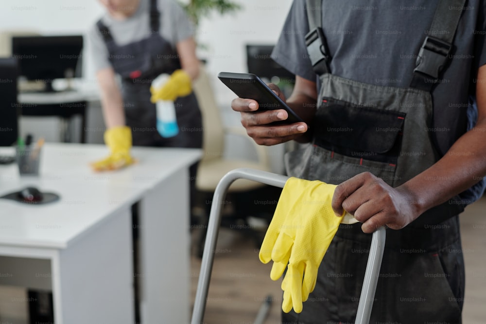 Manos de un joven limpiador masculino afroamericano de una empresa de servicios de limpieza moderna que elige música en el teléfono inteligente durante el trabajo