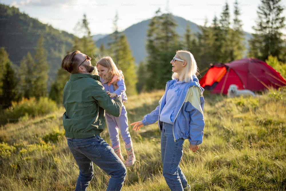 Young caucasian couple with little girl have fun while travel with tent in the mountains. Happy family summer vacation at campsite. Father tossing daughter in his arms