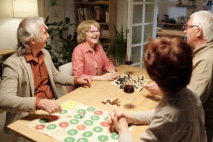 Cheerful aged woman laughing during name game with friends gathered by table with chessboard and paper with green and red circles