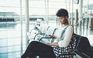 Portrait d’une jeune fille de touriste brésilienne douce en pull blanc, avec un sac à dos assis dans la salle d’attente du terminal de l’aéroport ou du dépôt de la gare, ou du centre commercial et de la messagerie à l’aide d’un smartphone