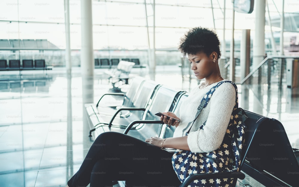 Portrait of young sweet Brazilian tourist girl in white pullover, with backpack sitting in waiting room of airport terminal or railway station depot, or shopping mall and messaging using smartphone
