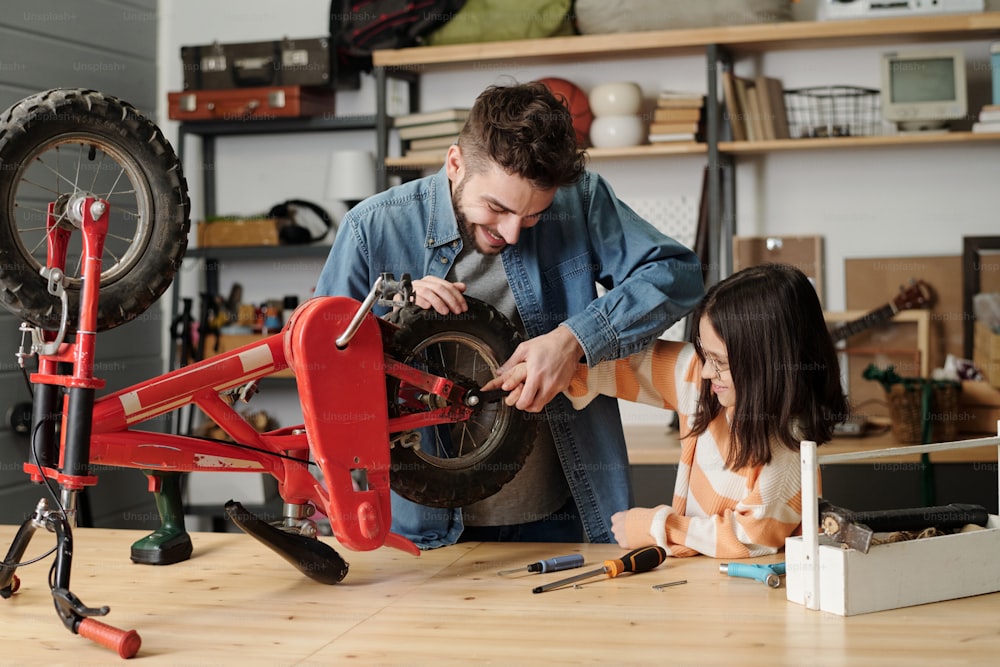Young man holding hand of his little daughter while checking wheels of her bicycle or repairing it by wooden table in garage