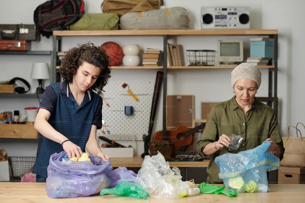 Grandmother and grandson sorting various kinds of waste into different cellophane sacks while standing by large table in garage