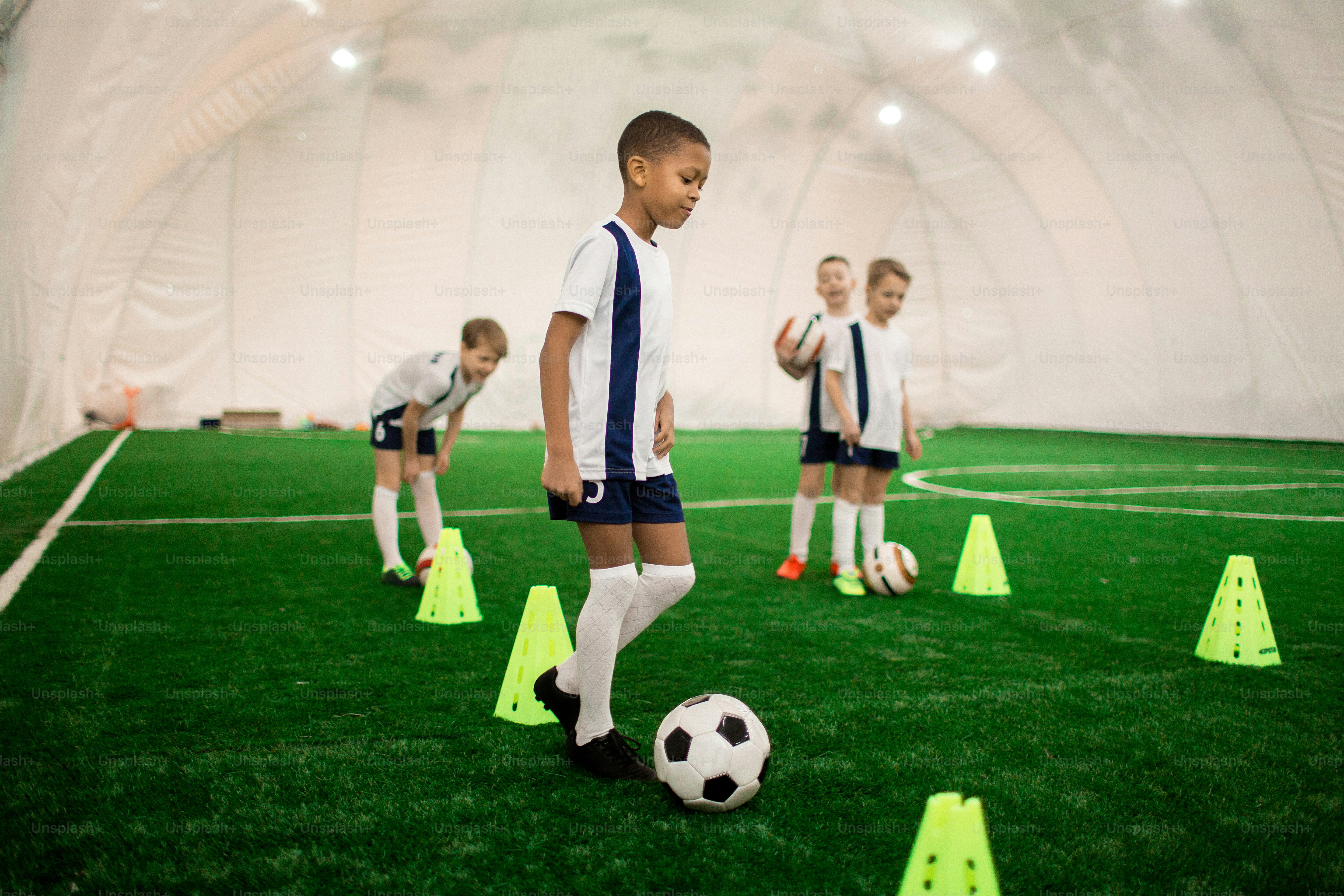 Salah satu anak laki-laki berseragam sepak bola berlatih dengan bola di lapangan hijau dengan kerucut