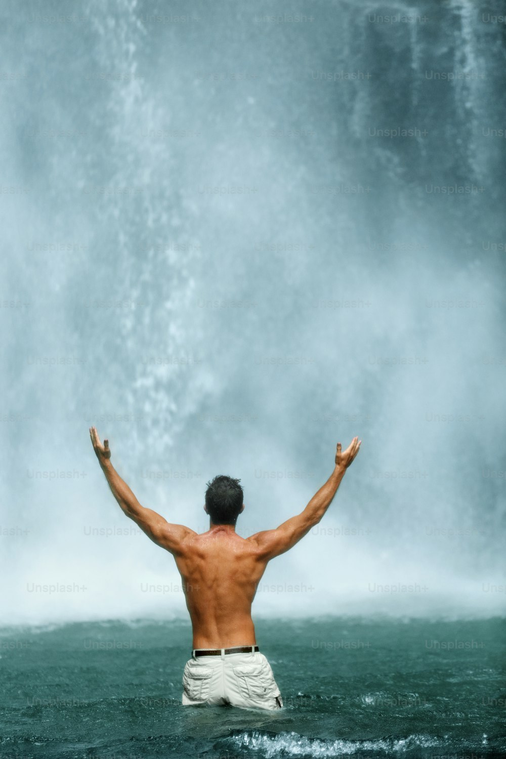 Free Man Enjoying Nature Beauty, Raising Hands Up To Sky. Male Relaxing, Standing In Water Near Beautiful Waterfall On Summer Travel Vacation. Freedom Of Life Concept.