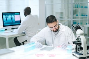 Junger seriöser männlicher Chemiker in Weißkittel, Schutzhandschuhen und Brillen, der Substanz in Petrischale nach Arbeitsplatz untersucht