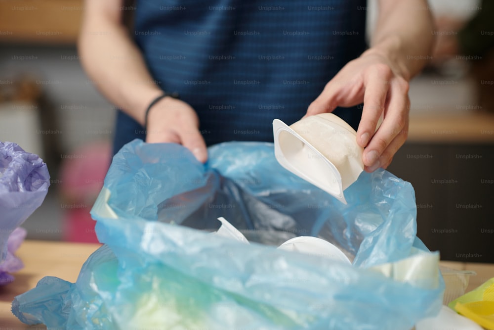 Mãos de um jovem cara colocando pequeno recipiente de comida de plástico branco no saco de celofane enquanto separa vários tipos de resíduos
