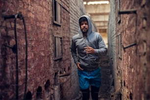 Portrait of focused motivated afro-american young handsome runner hooded man with earphones jogging inside of the abandoned place in the middle of two walls.