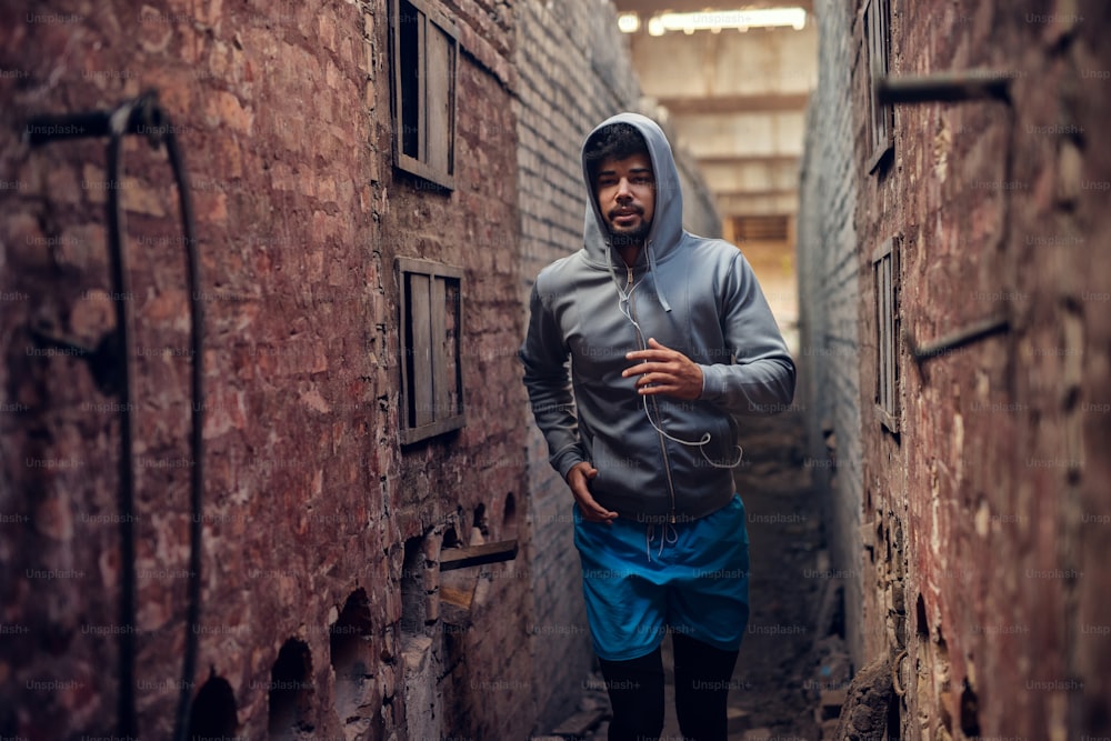 Portrait of focused motivated afro-american young handsome runner hooded man with earphones jogging inside of the abandoned place in the middle of two walls.