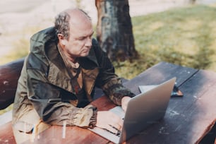 Aged partly bald man is sitting in a park with the laptop and digital tablet; adult male gamekeeper is sitting at the wooden table outdoors next to the tree and working on his netbook on a sunny day