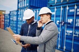 Engineer talking on a cellphone and reading an inventory list on a clipboard while standing with a colleague in a commercial shipping yard