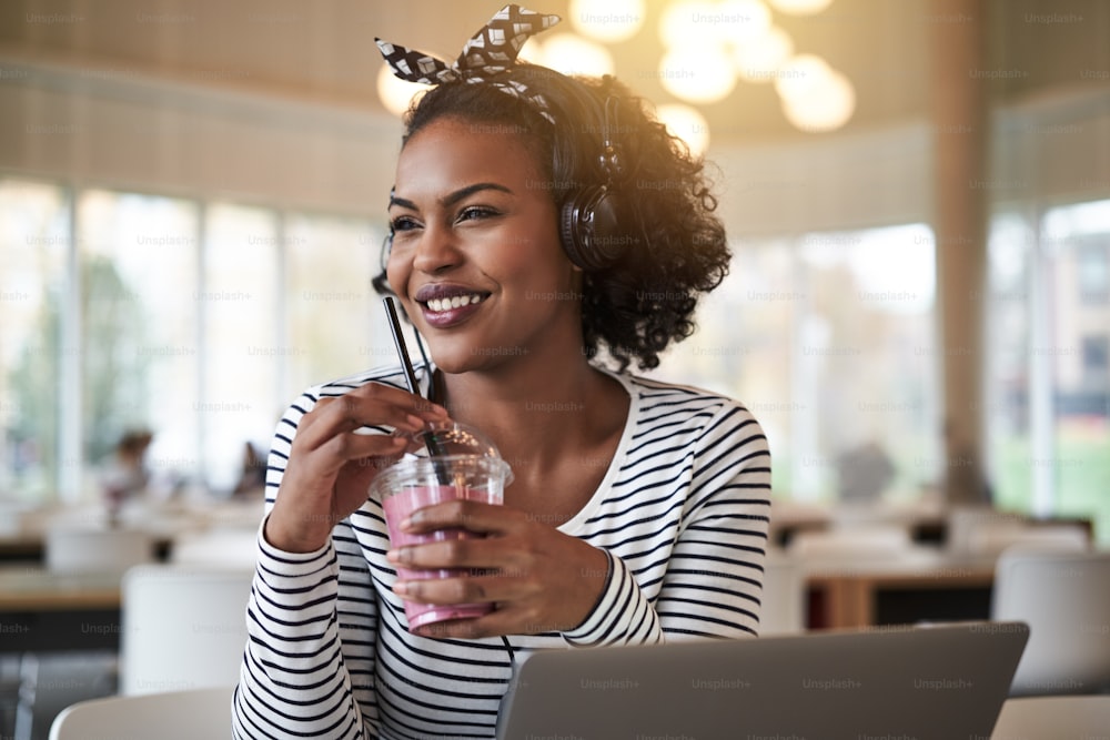 Junge afrikanische Studentin lächelt und arbeitet online, während sie auf dem Campus sitzt, einen Smoothie trinkt und Musik über Kopfhörer hört