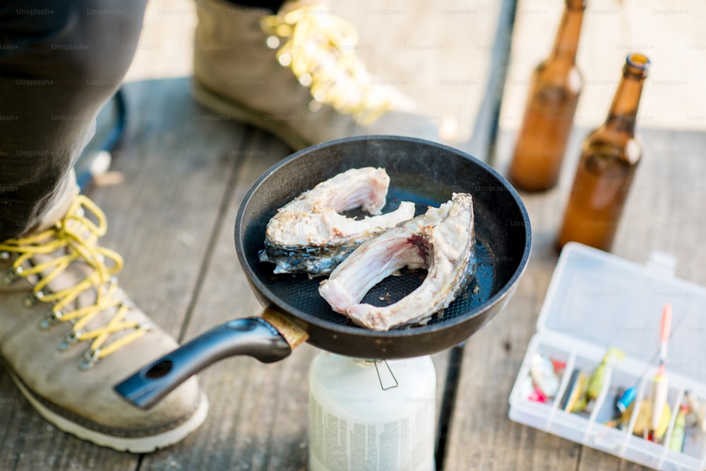 Frying two fish steakes on the burner during the picnic with beer and fishing tackles outdoors