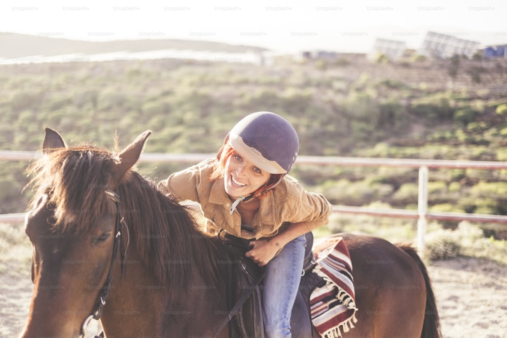 ride with a helmet for a beautiful attractive young woman with nice smile. jeans and casual clothes for outdoor girls in leisure activity with brown horse. animal therapy for alternative medicine