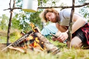 Young backpacker blowing on burning woods under pan with boiling water during trip in the forest