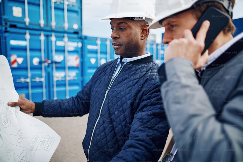 Zwei Ingenieure, die einen Bauplan in der Hand halten und auf einem Handy telefonieren, während sie in einer großen kommerziellen Werft an Schiffscontainern stehen
