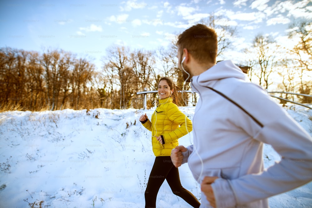 Young attractive healthy runner couple jogging with earphones on the snow covered road in nature in the morning.