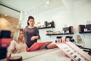 Young blonde woman looking at her reflection in a mirror while sitting in a salon chair looking through hair dye samples with her stylist