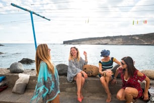 group of four young nice friends sitting speaking and laughing near the ocean. coast life in vacation leisure outdoor activity for fashion girls. enjoy the freedom and the lifestyle