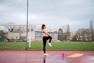 Seitenansicht der attraktiven Form junges Fitness-Mädchen, das sich streckt, während es das Knie draußen in der Nähe des Fußballplatzes hält.