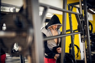 Close up portrait of focused and motivated muscular hooded man standing with earphones in the modern gym.