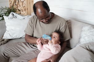 Vue en plongée d’un homme noir assis sur un canapé nourrissant son bébé au biberon avec du lait maternisé ou du lait maternel pompé
