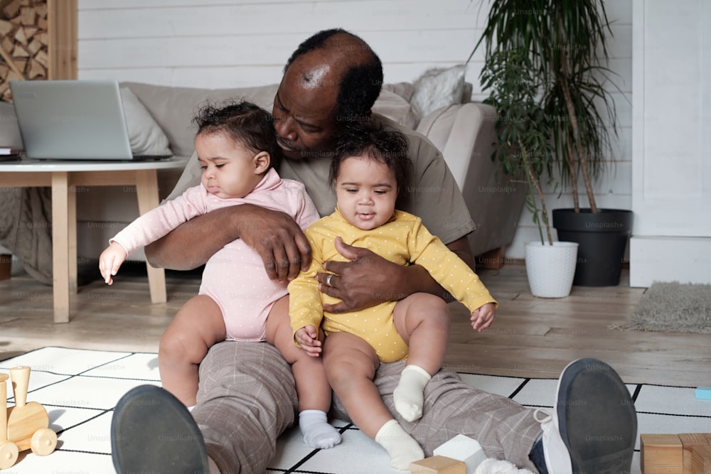 Glücklicher afroamerikanischer Vater, der zu Hause im gemütlichen Wohnzimmer auf dem Boden sitzt und seine Zwillingstöchter umarmt
