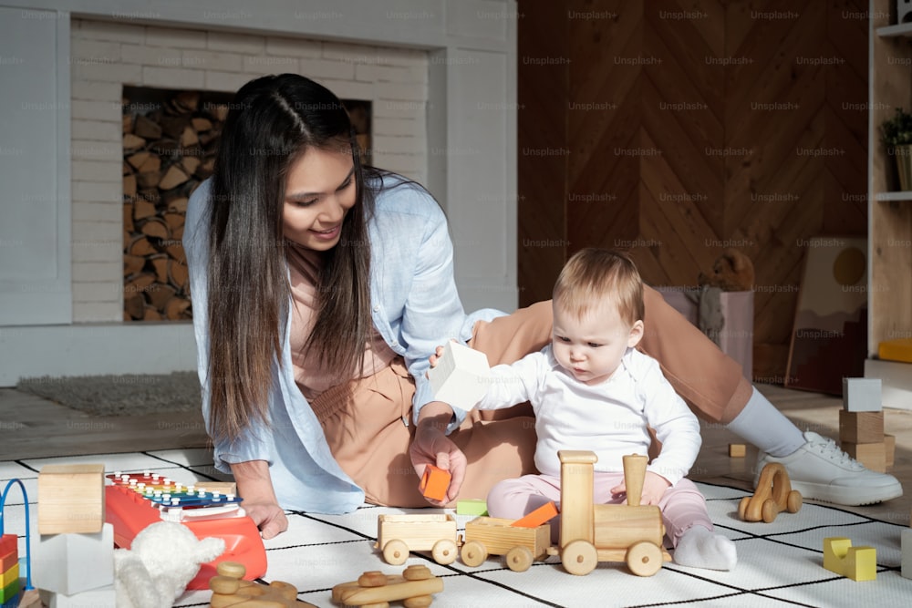 Liebevolle junge Mutter, die Zeit zu Hause verbringt und ihrer kleinen Tochter beim Spielen mit Holzspielzeug zusieht