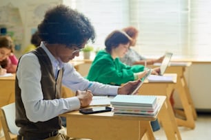 Side view of teenage guy with tablet carrying out individual assignment while sitting by desk against his classmates at lesson of anatomy