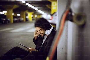 Close up de um elegante e brincalhão simplista skatista sentado na parede da garagem subterrânea em um terno e ouvindo música enquanto segurava um tablet.