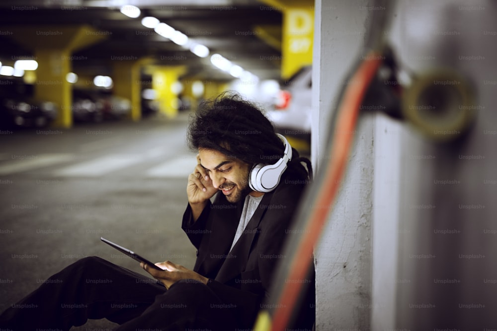 Close up of stylish playful smiling dreadlock skater man sitting leaned on the wall of the underground parking garage in a suit and listening music while holding a tablet.