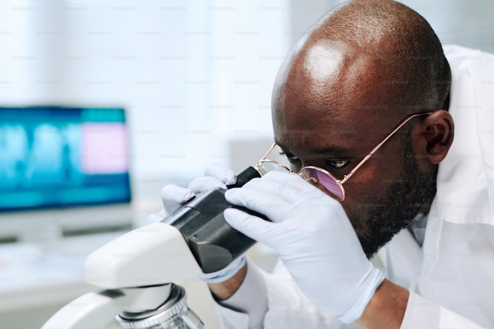 Kahlköpfiger afroamerikanischer Wissenschaftler in Handschuhen und Brillen betrachtet neue Viren oder Bakterien im Mikroskop, während er im Labor arbeitet