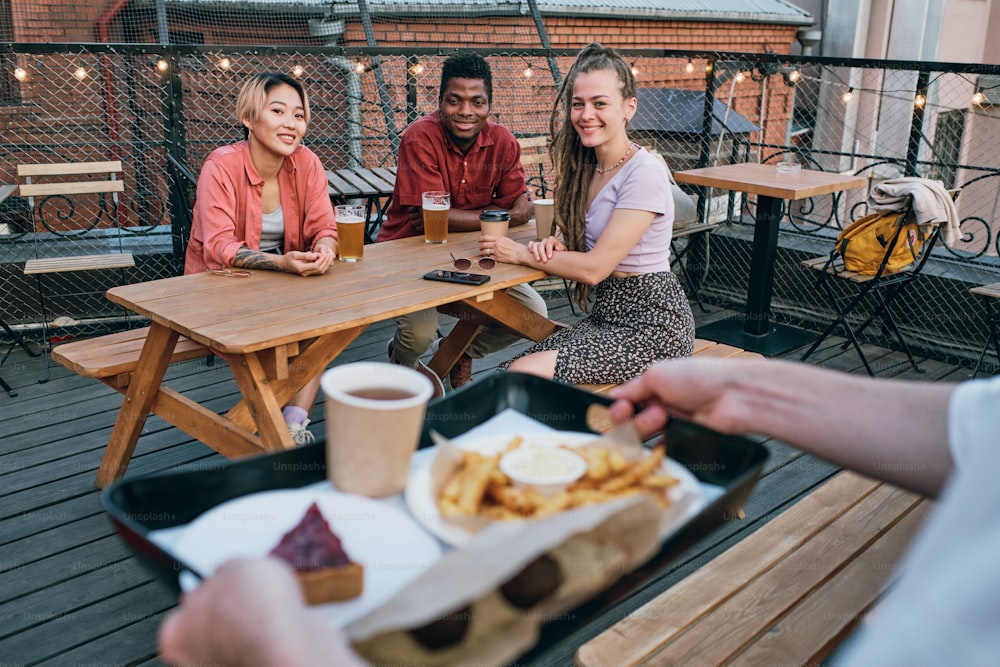 Tres jóvenes amigos interculturales felices que miran al camarero que lleva la bandeja con postre, comida rápida y bebida