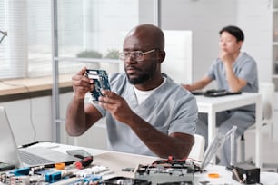 Joven reparador serio mirando la placa de circuito de un dispositivo electrónico en sus manos mientras está sentado en el lugar de trabajo contra una compañera de trabajo
