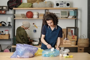Teenage guy sorting waste and putting it into two large cellophane sacks while standing by table in garage against his grandmother