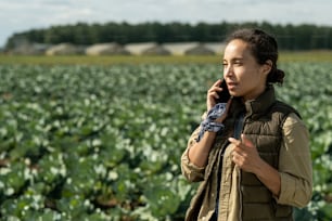 Young female farmer or owner of farm speaking on mobile phone against plantation