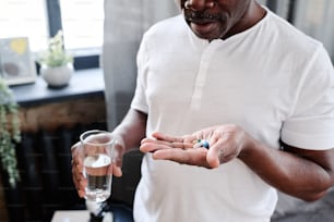 African American elderly male with glass of water holding pills on hand before taking them in the morning after sleep in bedroom