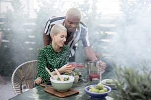 Black man taking juice for pouring in glass of his european girlfriend during dinner outdoors. Concept of relationship and enjoying time together. Modern domestic lifestyle. Woman sitting at table