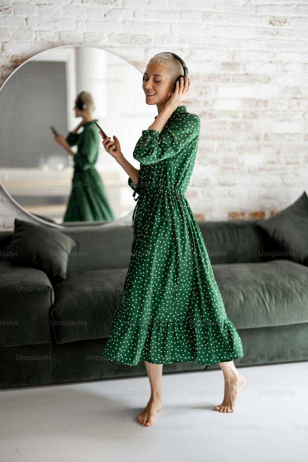 La mujer elegante con vestido verde disfruta de la música con auriculares y teléfono celular bailando en la sala de estar de casa